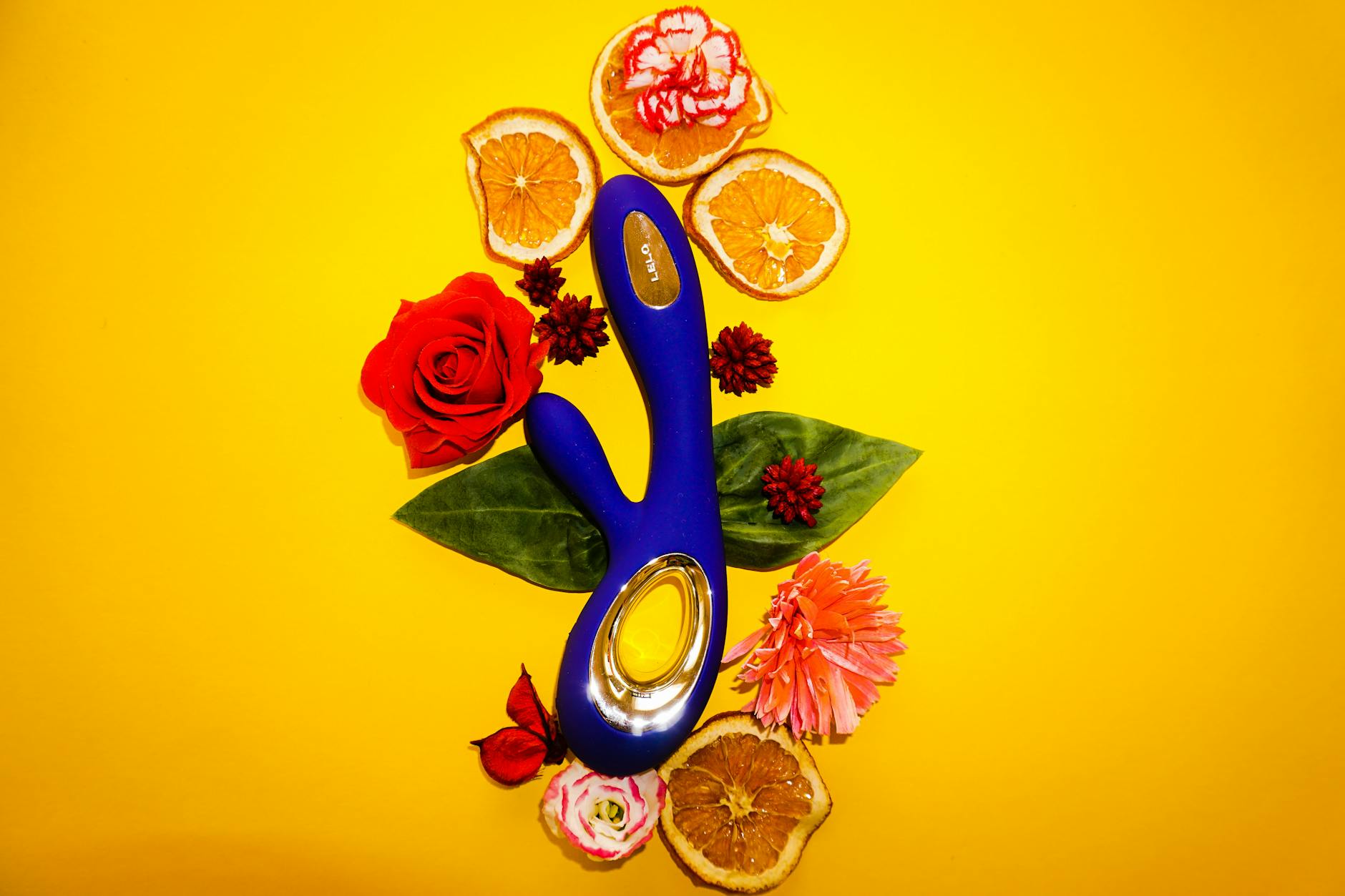 Blue vibrator surrounded by fresh citrus and flowers on a bright yellow surface.
