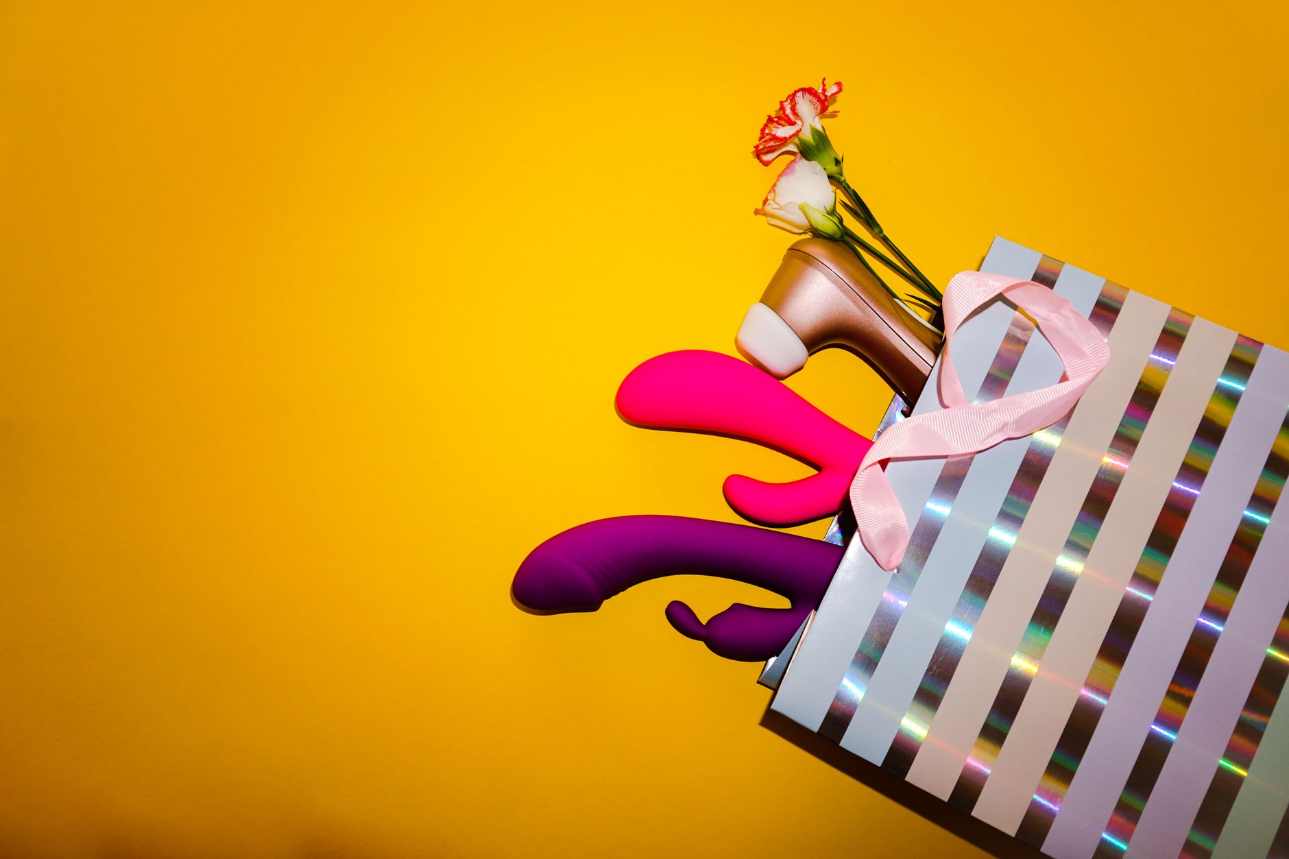 Colorful vibrators with flowers in a holographic gift bag on a bright yellow background